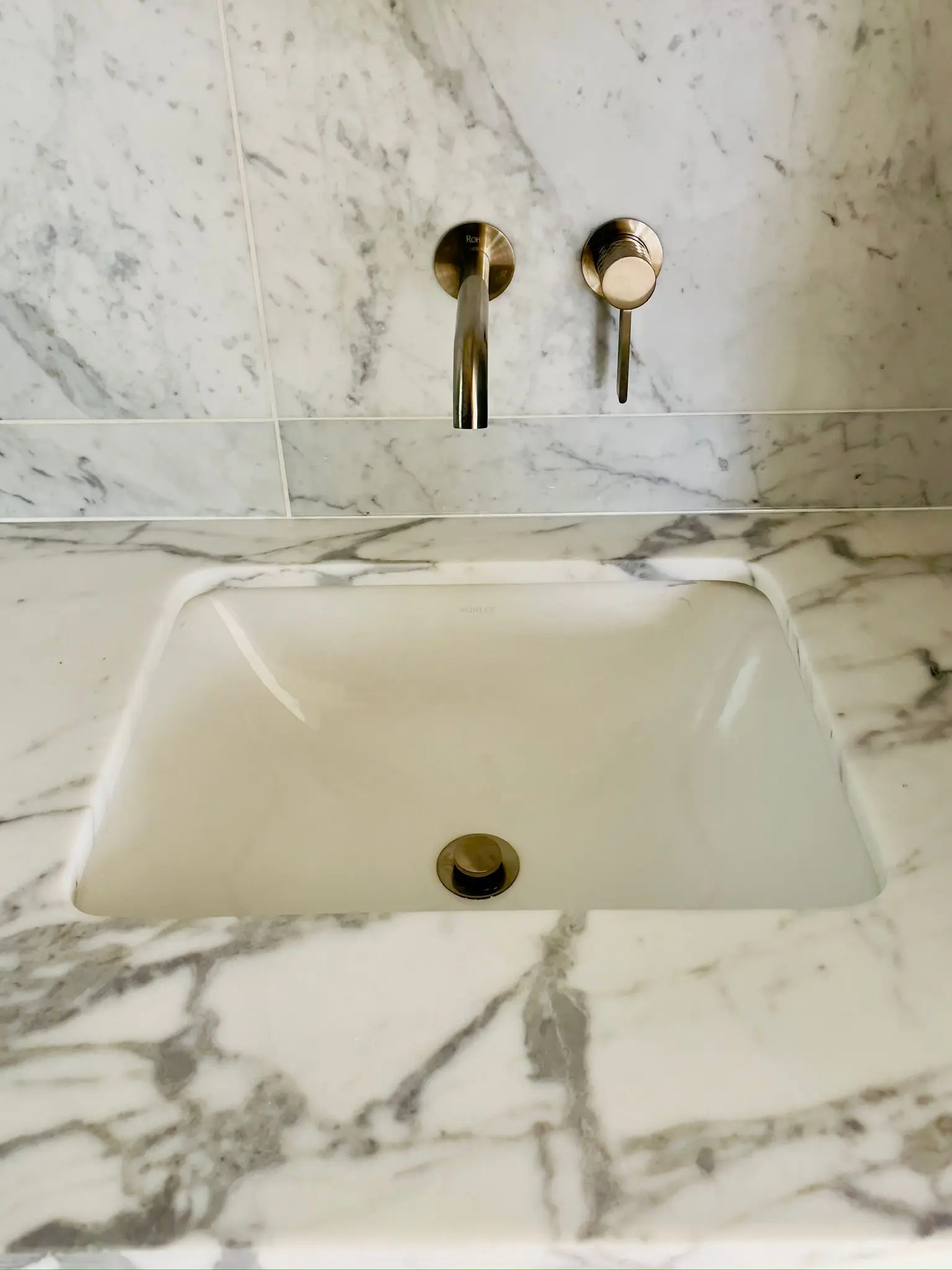 An undermount basin in marble, fed by a brass wall-mounted tap.