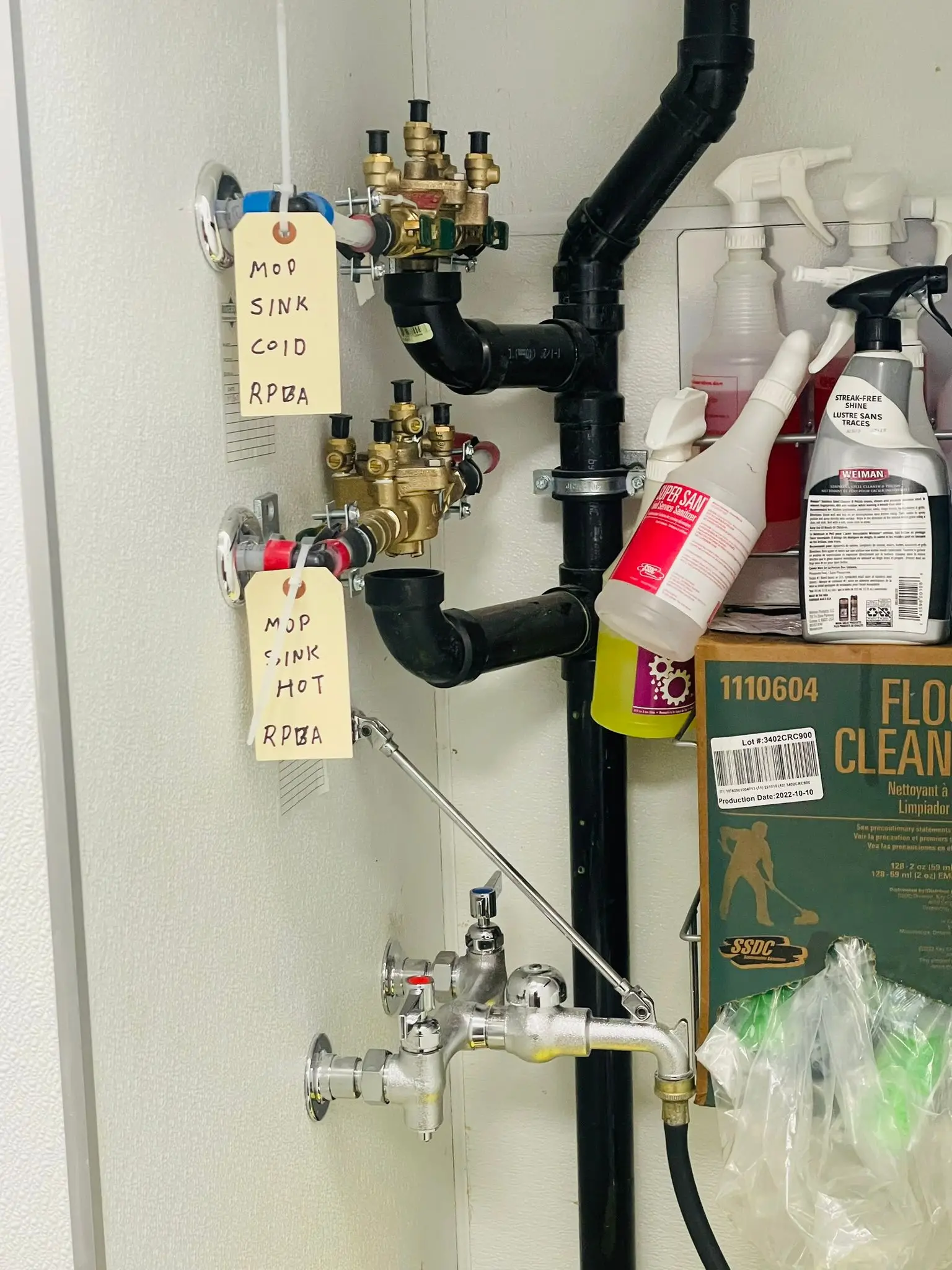 Labelled mop sink supplies and a backflow assembly on a kitchen wall.